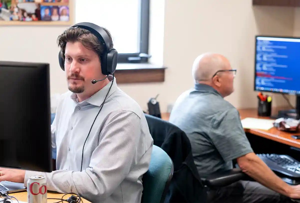 Two employees working at computers in an office, one wearing a headset.